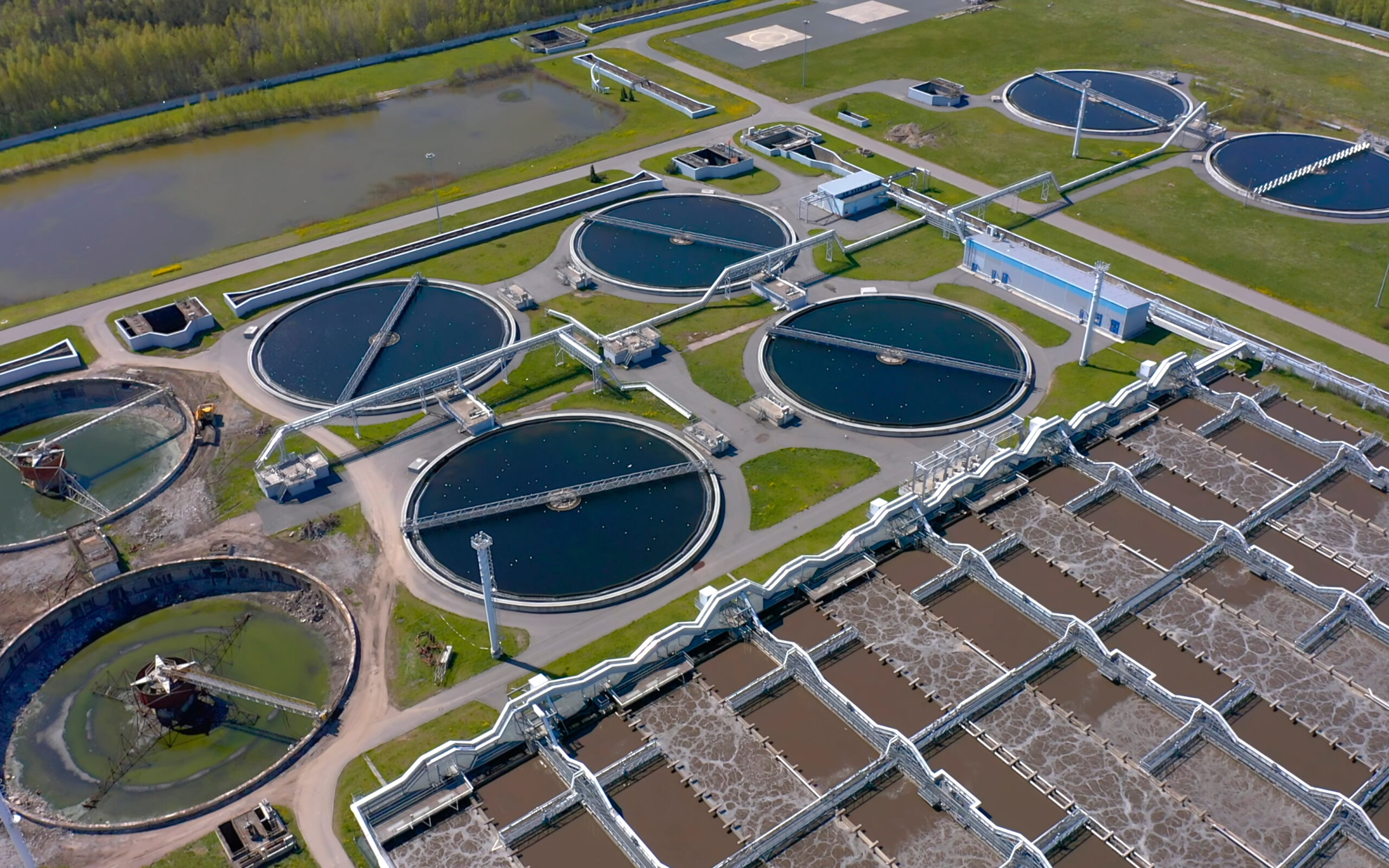 Vue aérienne d'une grande station d'épuration avec plusieurs bassins de décantation circulaires, canaux de traitement et zones végétalisées environnantes