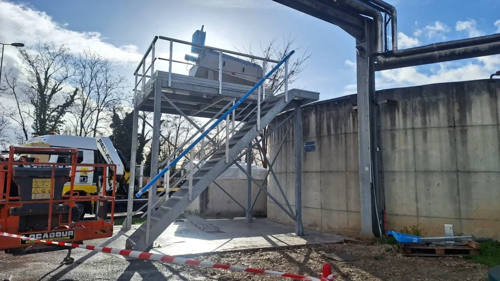Chantier de mise en service d'un dégrilleur BioScreen TRI Biotrade sur une plateforme métallique surélevée avec escalier d'accès, ouvrage béton et engin de chantier