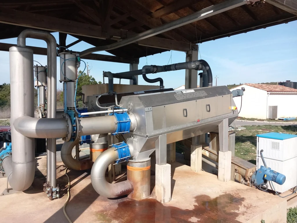 Dégrilleur BioScreen TRI Biotrade installé sous un abri métallique avec réseau de tuyauteries inox et raccords bleus sur une station d'épuration